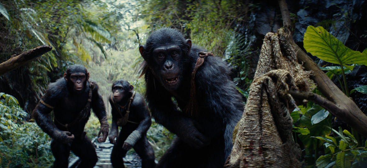 In a scene from the Kingdom of the Planet of the Apes, Noa (Owen Teague), Soona (Lydia Peckham) and Anaya (Travis Jeffery), three young apes, are depicted in a forest. They wear accessories from their village, the Eagle Clan, featuring knit details and patterns on their hands and around their chest. On the right side of a photo, a piece of straw-like fabric is placed over a branch. 