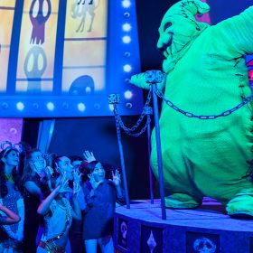 A life-size figure of Oogie Boogie, in fluorescent green, stands on a small stage on the righthand side of the image, from a previous Oogie Boogie Bash at Disneyland Resort. Behind Oogie Boogie is his familiar wheel of misfortune. In front of the stage are people attending the party, smiling and having a good time. Above the heads of the partygoers are iconic graphics from Tim Burton’s The Nightmare Before Christmas.