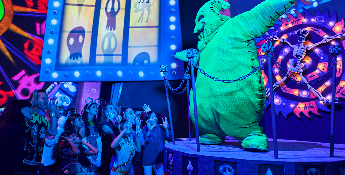 A life-size figure of Oogie Boogie, in fluorescent green, stands on a small stage on the righthand side of the image, from a previous Oogie Boogie Bash at Disneyland Resort. Behind Oogie Boogie is his familiar wheel of misfortune. In front of the stage are people attending the party, smiling and having a good time. Above the heads of the partygoers are iconic graphics from Tim Burton’s The Nightmare Before Christmas.