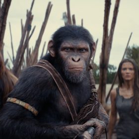 In a scene from Kingdom of the Planet of the Apes, the chimpanzee Noa (played by Owen Teague) stands in a fierce posture, facing the camera with a weapon of some kind at ready, held in front of his chest. Behind him to the left is the orangutan Raka (played by Peter Macon); to the right behind him is the female human Nova (played by Freya Allan). They appear to be on a beach, standing near a barrier made of sharpened sticks and nets.