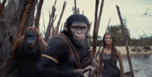 In a scene from Kingdom of the Planet of the Apes, the chimpanzee Noa (played by Owen Teague) stands in a fierce posture, facing the camera with a weapon of some kind at ready, held in front of his chest. Behind him to the left is the orangutan Raka (played by Peter Macon); to the right behind him is the female human Nova (played by Freya Allan). They appear to be on a beach, standing near a barrier made of sharpened sticks and nets.