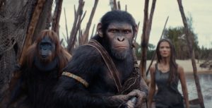 In a scene from the Kingdom of the Planet of the Apes, Anaya (Travis Jeffrey), Noa (Owen Teague), and Soona (Lydia Peckham), three young apes, are depicted reading a book. Anaya (left) holds a torch, adding light to the dimly-lit frame.