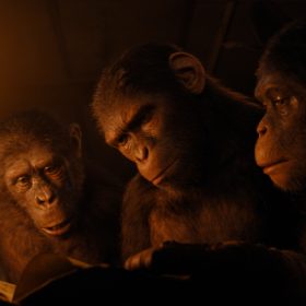 In a scene from the Kingdom of the Planet of the Apes, Anaya (Travis Jeffrey), Noa (Owen Teague), and Soona (Lydia Peckham), three young apes, are depicted reading a book. Anaya (left) holds a torch, adding light to the dimly-lit frame.