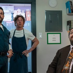 In a scene from Season 3 of The Bear, pictured from left to right are Theodore Fak (played by Ricky Staffieri), Carmen “Carmy” Berzatto (played by Jeremy Allen White) and Neil Fak (played by Matty Matheson). They are standing in backroom of a restaurant with a row of lockers behind them. They face forward, toward the camera. Ted Fak, wearing a blue-green handyman’s uniform stands, and points with his left hand toward something above eye level in the distance, unseen. Carmy, in a white T-shirt with navy blue kitchen apron stands and looks up. Neil, seated at a desk with his chair facing outward and wearing a brown suit with a floral and brown decorated tie, looks up at it too. 