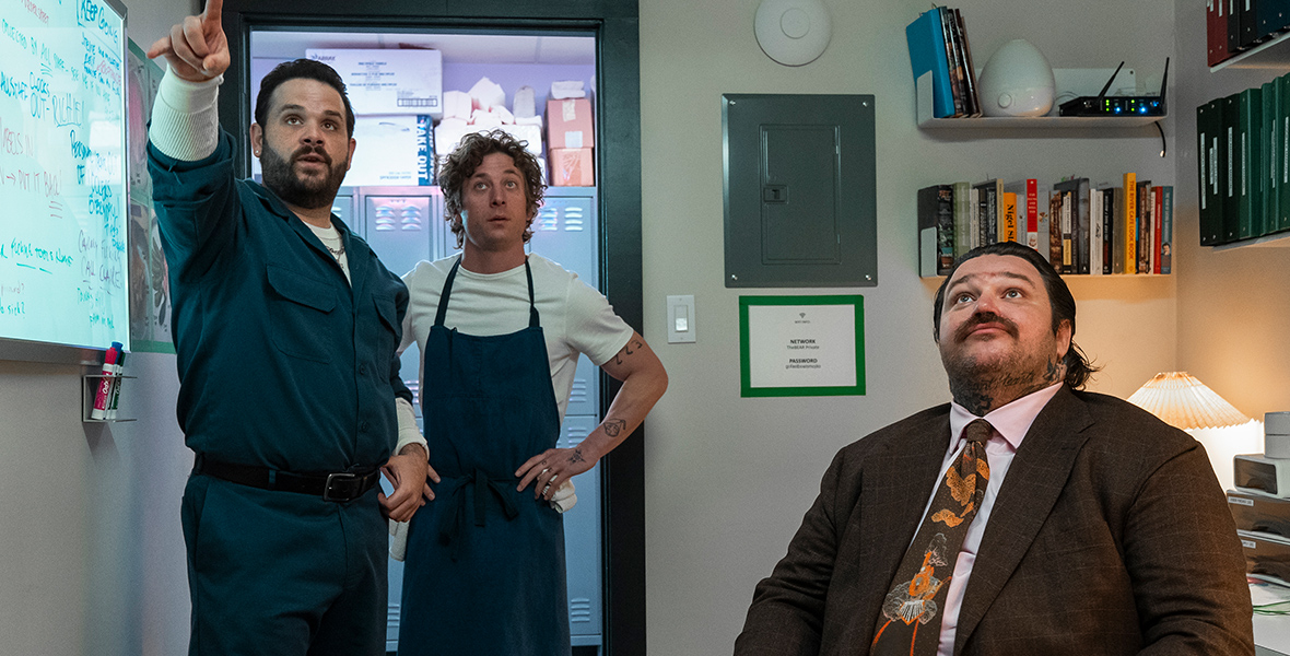 In a scene from Season 3 of The Bear, pictured from left to right are Theodore Fak (played by Ricky Staffieri), Carmen “Carmy” Berzatto (played by Jeremy Allen White) and Neil Fak (played by Matty Matheson). They are standing in backroom of a restaurant with a row of lockers behind them. They face forward, toward the camera. Ted Fak, wearing a blue-green handyman’s uniform stands, and points with his left hand toward something above eye level in the distance, unseen. Carmy, in a white T-shirt with navy blue kitchen apron stands and looks up. Neil, seated at a desk with his chair facing outward and wearing a brown suit with a floral and brown decorated tie, looks up at it too. 