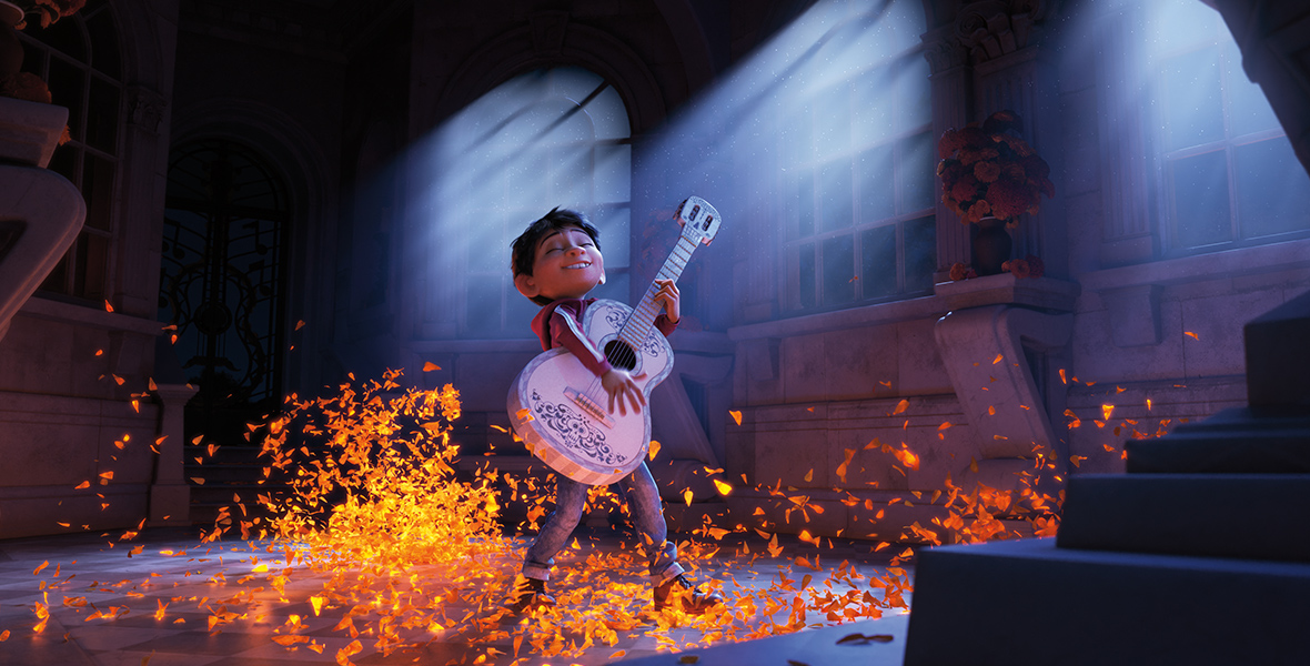 In a scene from the movie Coco, Miguel (voiced by Anthony Gonzalez) strums his guitar in a moonlit room. Glowing, orange petals gather on the floor and swirl around Miguel, who is wearing a red hoodie with white stripes along the sleeves, blue jeans, and black boots.