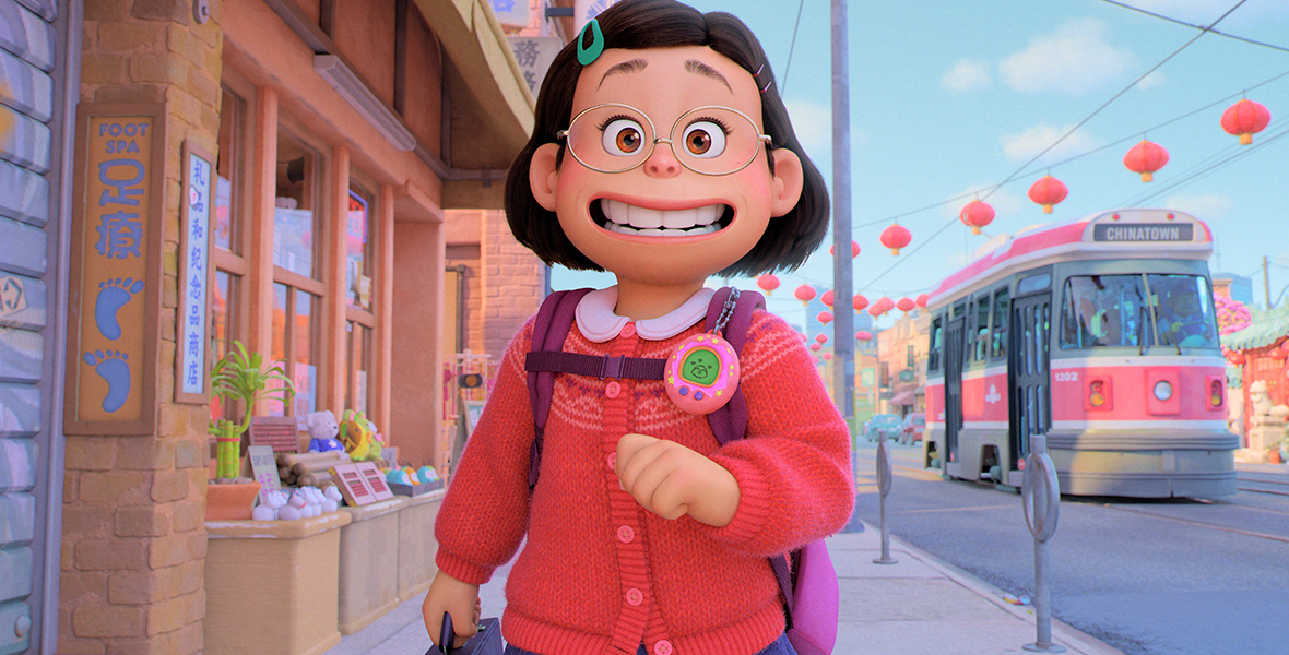 In a scene from Turning Red, Meilin Lee (Mei), (voiced by Rosalie Chian), strolls along the sidewalk. Her short, dark hair is adorned with colorful pins. Mei wears thin, oval glasses, and a red knit cardigan paired with a blue denim skirt. A Tamagotchi dangles from her pink backpack as she holds a briefcase in her right hand. Behind her, Toronto’s Chinatown reveals buildings, a bus, and the exterior of a foot spa.