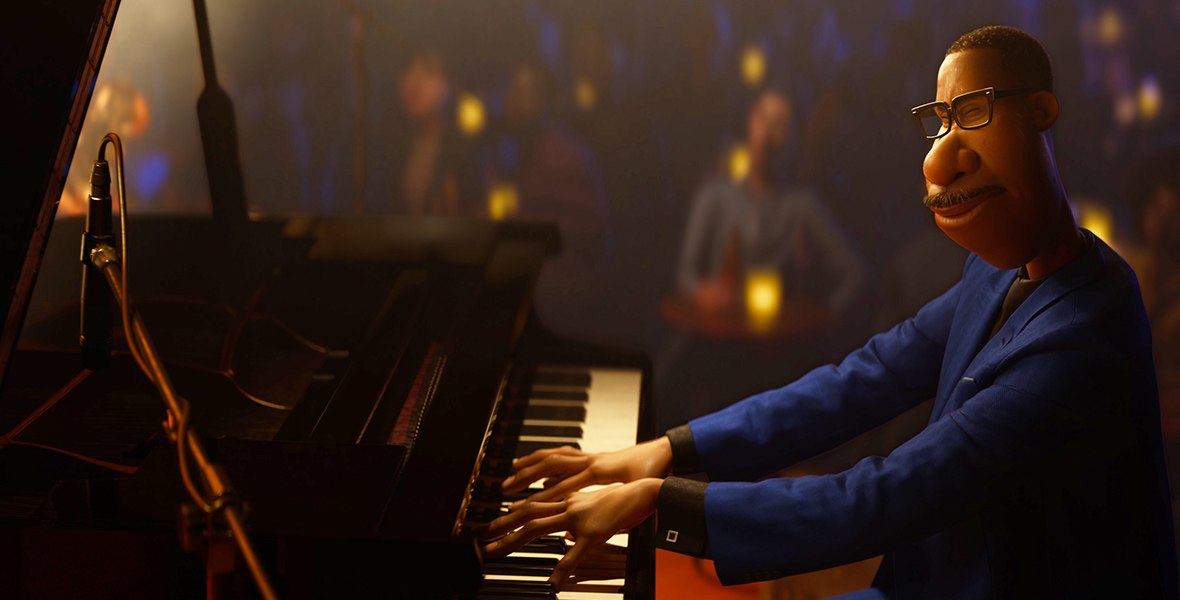 In a scene from Soul, Joe Gardner (voiced by Jamie Foxx) is playing the piano. He is wearing a blue suit, a brown shirt, and black square glasses. The background of the photo, though out of focus, depicts Joe’s audience.