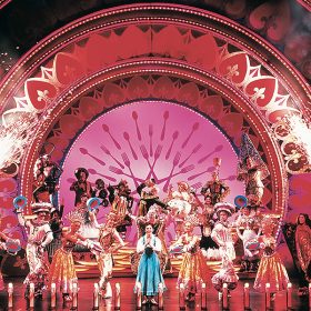 In a photo by Joan Marcus, the original Broadway cast of Disney’s Beauty and the Beast, including Susan Egan as Belle, performs the number “Be Our Guest.” Cast members are costumed as various pieces of cutlery or other household objects, and large champagne bottles flank the proscenium and are seen shooting firework-type sparkles in the air. On the apron of the stage are lamps that resemble candles.