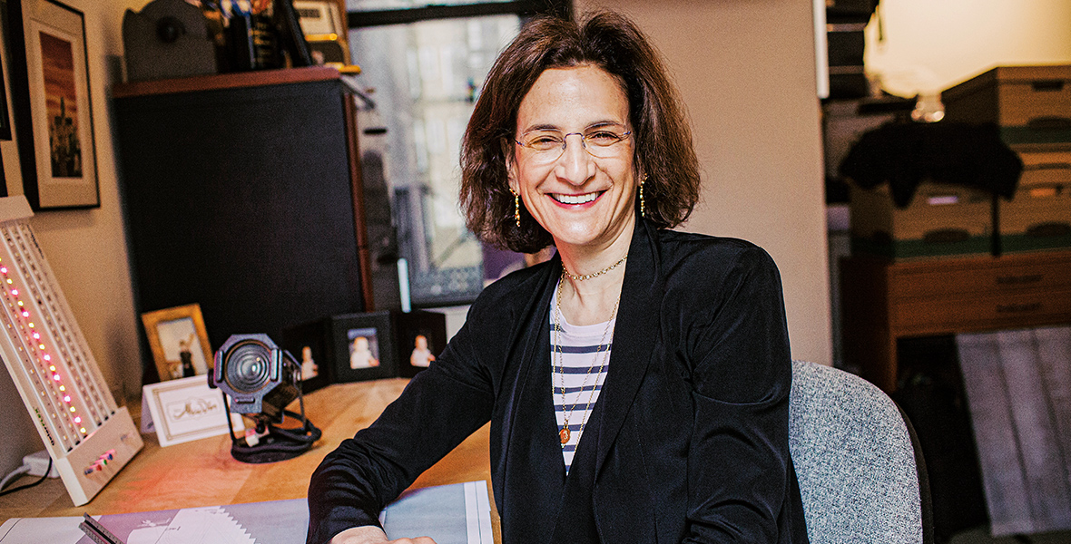 In an image by Jacqueline Harriet, Tony Award-winning lighting designer Natasha Katz is seen sitting at her desk. She is wearing glasses, as well as a black blazer with a black and white striped shirt underneath. A window can be seen in the background, and various design accoutrement are seen on her desk to the left.