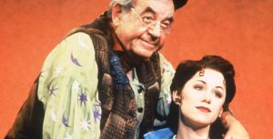 In an image by Joan Marcus, original Beauty and the Beast on Broadway cast member Susan Egan, as Belle, stands with fellow cast member Tom Bosley as Maurice. Bosley is standing behind Egan; he is wearing a hat, glasses, and a vest, and she is wearing Belle’s signature blue dress with a white apron.