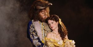 In an image by Joan Marcus, original Beauty and the Beast on Broadway cast member Susan Egan, as Belle, stands with fellow cast member Terrance Mann as the Beast. They are seen in their ornate “ballroom scene” costumes, including Belle’s iconic gold gown and the Beast’s blue and gold waistcoat. Mann behind Egan and looking down at her, while she is looking off to the left.