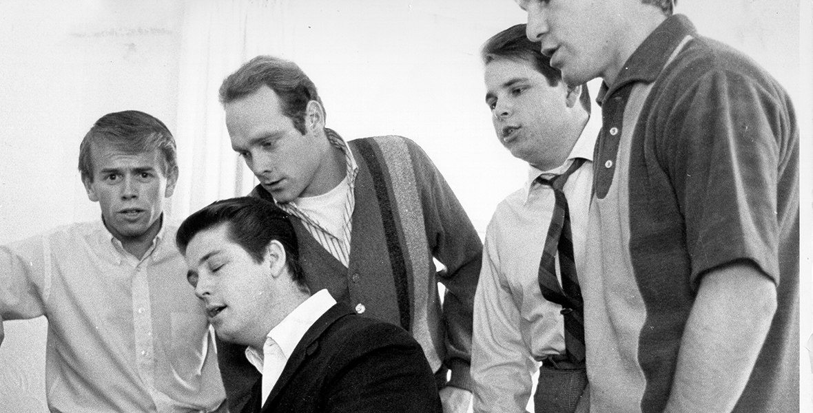 In a vintage black and white photo, Brian Wilson sits at a piano, immersed in song with his eyes closed as he plays. Standing beside him from left to right are the other members of The Beach Boys: Al Jardine, Mike Love, Carl Wilson, and Dennis Wilson, who are singing along.