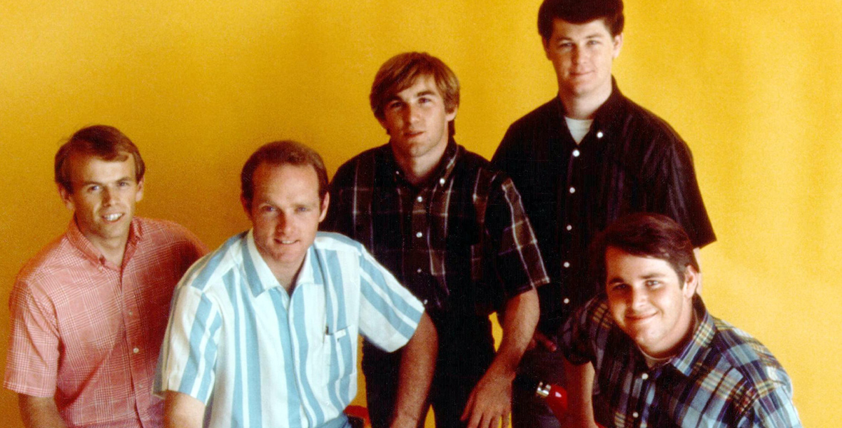 The Beach Boys pose for a portrait against a yellow backdrop. From left to right are Al Jardine, Mike Love, Dennis Wilson, Brian Wilson, and Carl Wilson. They are all wearing short sleeve plaid shirts, pants and white loafers. Love and Carl Wilson are sitting on two red mopeds. 