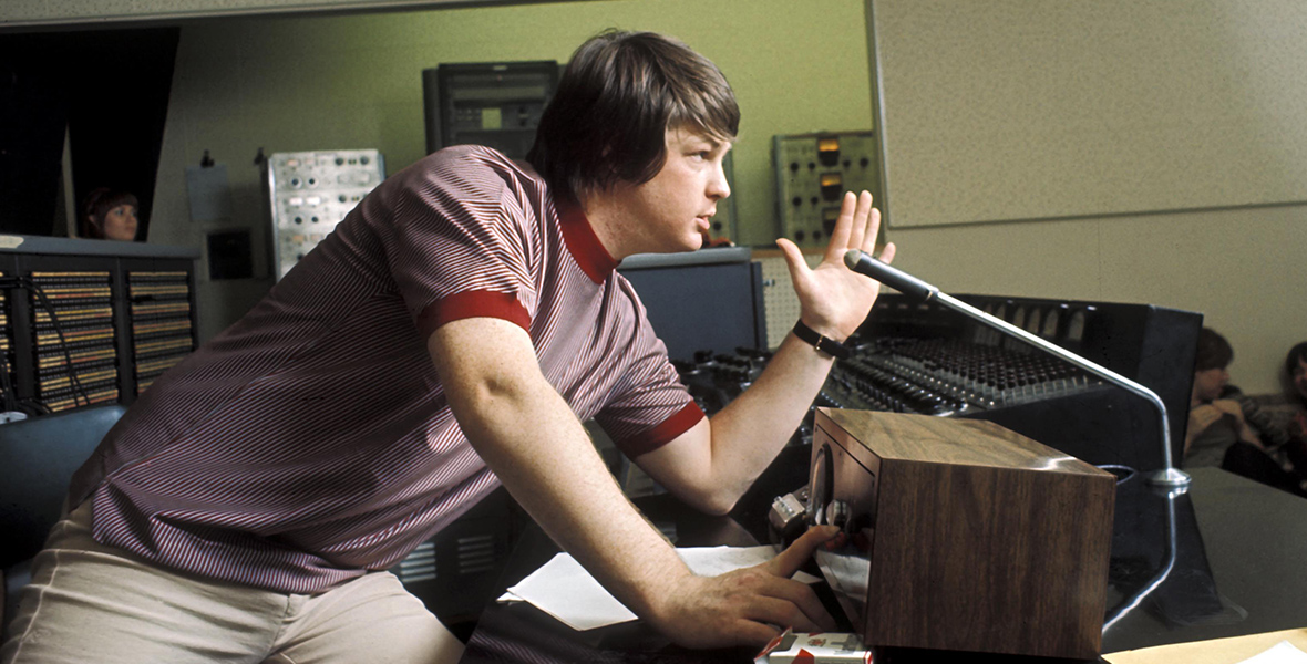 Brian Wilson from the band The Beach Boys is leaning on a desk in a recording studio control room. He is wearing a red and white striped t-shirt and beige pants. He gazes straight ahead, surrounded by recording equipment in the background.