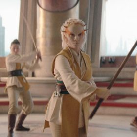 In a scene from The Acolyte, Jedi Padawan Jecki Lon (Dafne Keen) is wearing a cream-colored Jedi robe with a brown belt and holding a brown spear. She has orange paint on the sides of her head and small thorns along the perimeter of her forehead. Two Jedi are behind her to her left, and two more are behind her to her right. The interior space is circular, with the Jedi in the center and red and cream stairs surrounding them. Outside the building, skyscrapers are visible.