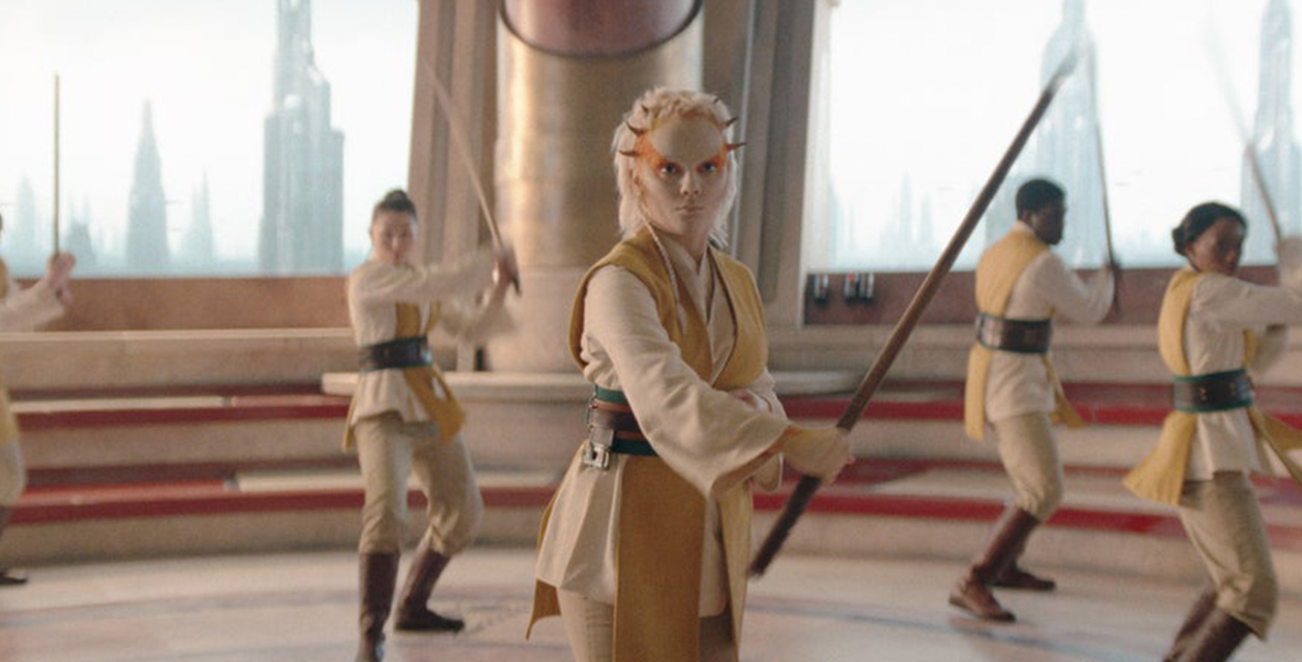 In a scene from The Acolyte, Jedi Padawan Jecki Lon (Dafne Keen) is wearing a cream-colored Jedi robe with a brown belt and holding a brown spear. She has orange paint on the sides of her head and small thorns along the perimeter of her forehead. Two Jedi are behind her to her left, and two more are behind her to her right. The interior space is circular, with the Jedi in the center and red and cream stairs surrounding them. Outside the building, skyscrapers are visible.