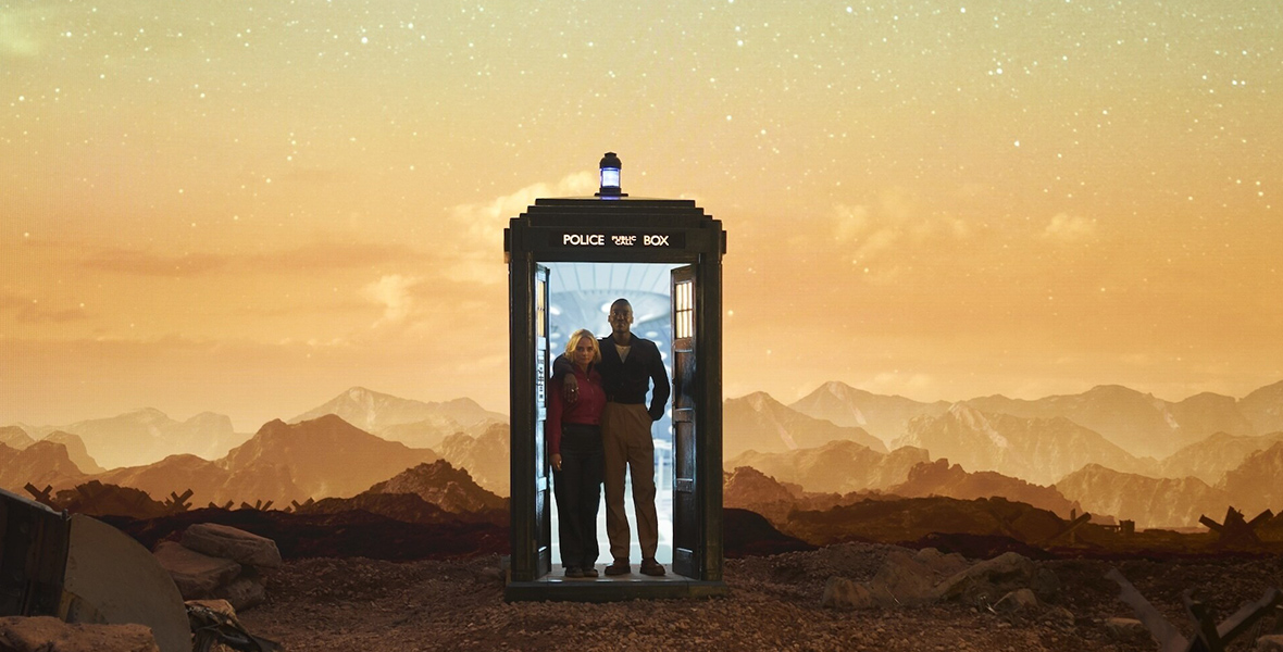 In a scene from Doctor Who, The Doctor (Ncuti Gatwa) and Ruby Sunday (Millie Gibson) are standing at the edge of the TARDIS, a police phone box. In the background are mountain peaks and a starry sky with a yellow hue. The TARDIS is on dry, rocky ground.