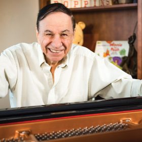 Richard M. Sherman sits behind a piano and smiles in Walt Disney's office.