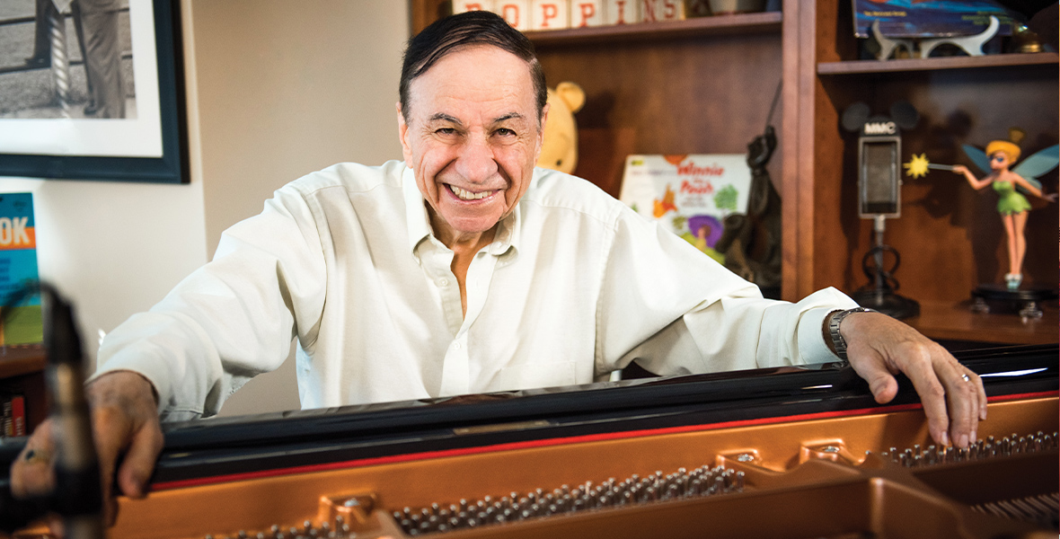 Richard M. Sherman sits behind a piano and smiles in Walt Disney's office.