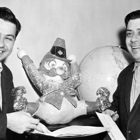 IMAGE00 ALT TEXT: In a black-and-white vintage photo, Disney Legends Richard M. Sherman (left) and Robert B. Sherman (right) are leaning on a black stool with a toy clown on it. Both Sherman brothers are looking to their right and smiling, dressed in dark-colored suits and light-colored shirts.