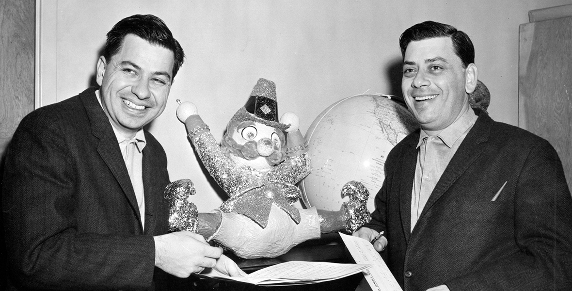 IMAGE00 ALT TEXT: In a black-and-white vintage photo, Disney Legends Richard M. Sherman (left) and Robert B. Sherman (right) are leaning on a black stool with a toy clown on it. Both Sherman brothers are looking to their right and smiling, dressed in dark-colored suits and light-colored shirts.