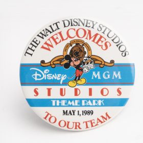 The Grand Opening promotional button is a white and blue circle button with text that reads “The Walt Disney Studios Welcomes Disney-MGM Studios Theme Park to Our Team, May 1, 1989.” Mickey Mouse holds a clapboard in the center of the button, embedded in the Disney-MGM Studios logo.