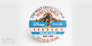 The Grand Opening promotional button is a white and blue circle button with text that reads “The Walt Disney Studios Welcomes Disney-MGM Studios Theme Park to Our Team, May 1, 1989.” Mickey Mouse holds a clapboard in the center of the button, embedded in the Disney-MGM Studios logo.