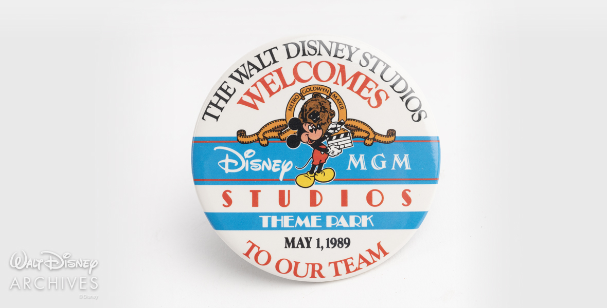 The Grand Opening promotional button is a white and blue circle button with text that reads “The Walt Disney Studios Welcomes Disney-MGM Studios Theme Park to Our Team, May 1, 1989.” Mickey Mouse holds a clapboard in the center of the button, embedded in the Disney-MGM Studios logo.
