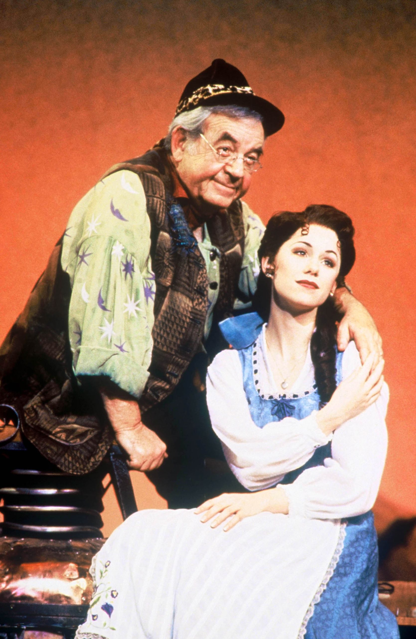 In an image by Joan Marcus, original Beauty and the Beast on Broadway cast member Susan Egan, as Belle, stands with fellow cast member Tom Bosley as Maurice. Bosley is standing behind Egan; he is wearing a hat, glasses, and a vest, and she is wearing Belle’s signature blue dress with a white apron.