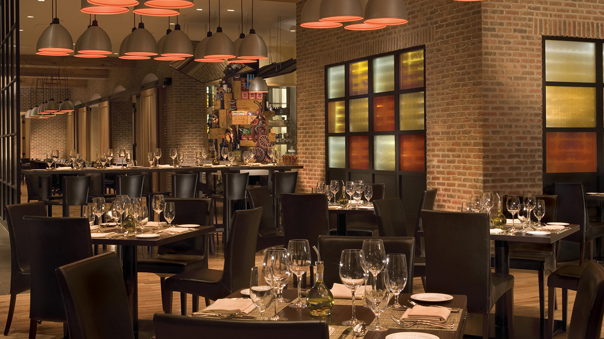 We are looking at a wonderful view of the inside of the high-end dining Il Mulino restaurant, located in the Dolphin Hotel at Walt Disney World. Empty wooden square tables have been set for the dinner rush. This includes water/wine glasses, silver utensils, napkins, and plates. In the center of each table sits a bottle of olive oil. The walls are made of orange brick while some areas have multicolored frosted windows.