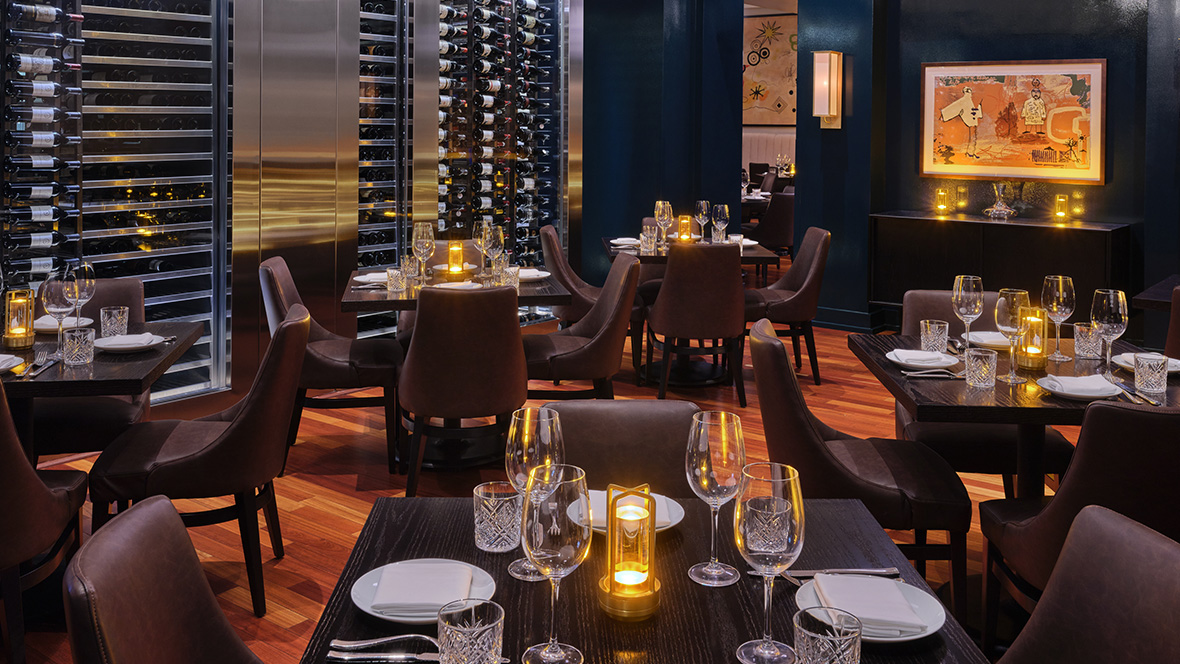 We are looking at a wonderful view of the inside of the high-end dining Il Mulino restaurant, located in the Dolphin Hotel at Walt Disney World. Empty wooden square tables have been set for the dinner rush. This includes water/wine glasses, silver utensils, napkins, and plates. In the center of each table sits a bottle of olive oil. The walls are made of orange brick while some areas have multicolored frosted windows. ALT TEXT (Michael_Minas_Bourbon_Steak1180664): We are looking at a photo of the inside of Bourbon Steak Orlando, a Michael Mina Restaurant located in the Walt Disney World Dolphin Hotel. From the photo, we can immediately tell that this is a fine-dining establishment. Wine glasses line the racks on silver shelves towards the back of the room. The flooring is a light wood, and each empty square table has been set for dinner. Water glasses, wine glasses, plates, napkins, and utensils have been set, and in the center of each table is a lit candle. The back wall is a rich blue color with art scattered throughout.