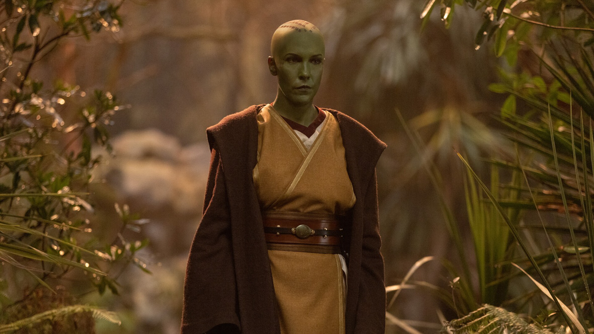 In an image from Star Wars: The Acolyte, Jedi Master Vernestra Rwoh, a green humanoid alien, stands in a forest looking at something offscreen.