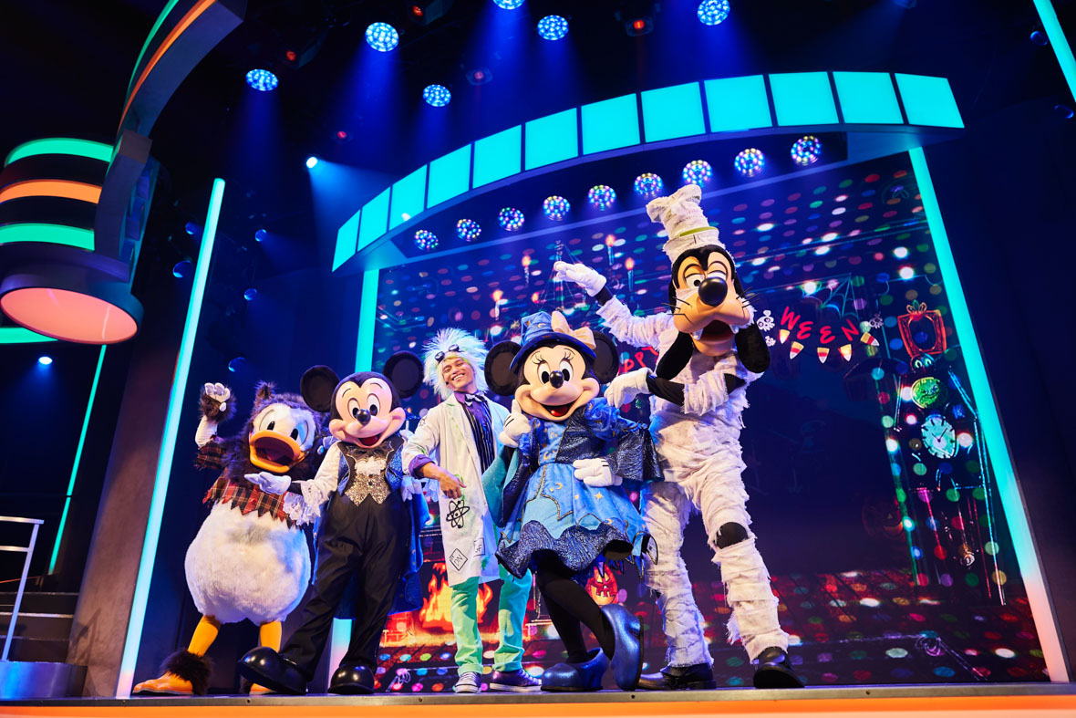 From left to right: Donald Duck, Mickey Mouse, a cast member dressed as a scientist, Minnie Mouse, and Pluto are performing on stage during the Mickey’s Trick and Treat daytime show. Donald is wearing a hairy brown headpiece and a cropped plaid top; Mickey is wearing a tuxedo with a bat-shaped bowtie; Minnie is wearing a blue wizard hat and dress; and Pluto is dressed as a mummy. The stage is filled with neon lights and Halloween decorations, such as candy corn.