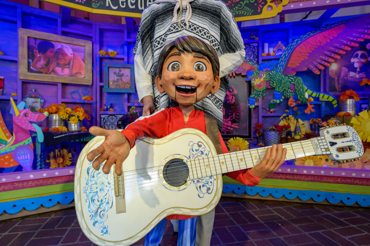 A life-size statue of Miguel from Disney and Pixar’s Coco holds a white guitar adorned with blue patterns in his left hand, while extending his right hand toward the camera. He wears a red zip-up jacket with white stripes and blue jeans. Behind him, decorations feature memorabilia of items from the film, such as a photo of Miguel with his grandmother and Aztec marigolds in yellow, orange, and red.