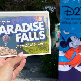 A hand holds up a postcard with text that reads “Fly up to Paradise Falls” and “A land lost in time.” Carl’s balloon cart is in the background of a grassy landscape and blue sky.