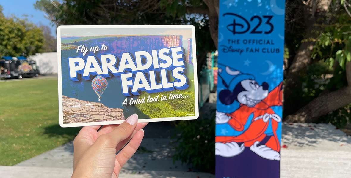 A hand holds up a postcard with text that reads “Fly up to Paradise Falls” and “A land lost in time.” Carl’s balloon cart is in the background of a grassy landscape and blue sky.