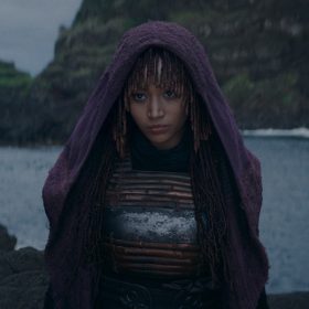 In an image from Star Wars: The Acolyte, Mae, a cloaked human, stands on alongside a body of water, staring intently at something offscreen.
