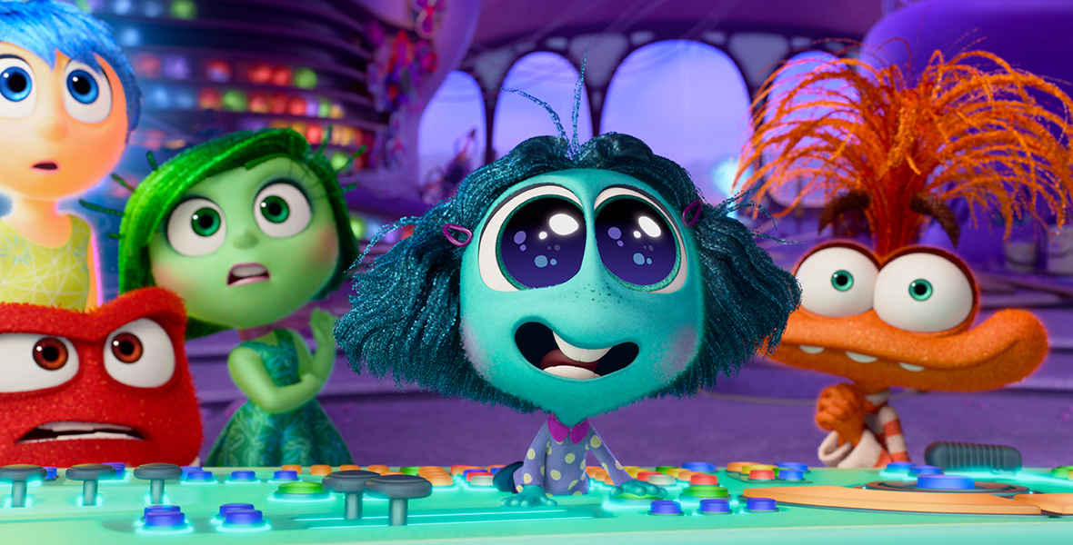 In a scene from Inside Out 2, Joy (voiced by Amy Poehler), Anger (voiced by Lewis Black), Disgust (voiced by Liza Lapira), Envy (voiced by Ayo Edebiri), and Anxiety (voiced by Maya Hawke) stand behind Riley’s Headquarters console, now teal, reflecting Envy’s influence. Envy lies atop the console. Joy, Anger, and Disgust wear shocked expressions, while Anxiety appears pleased.