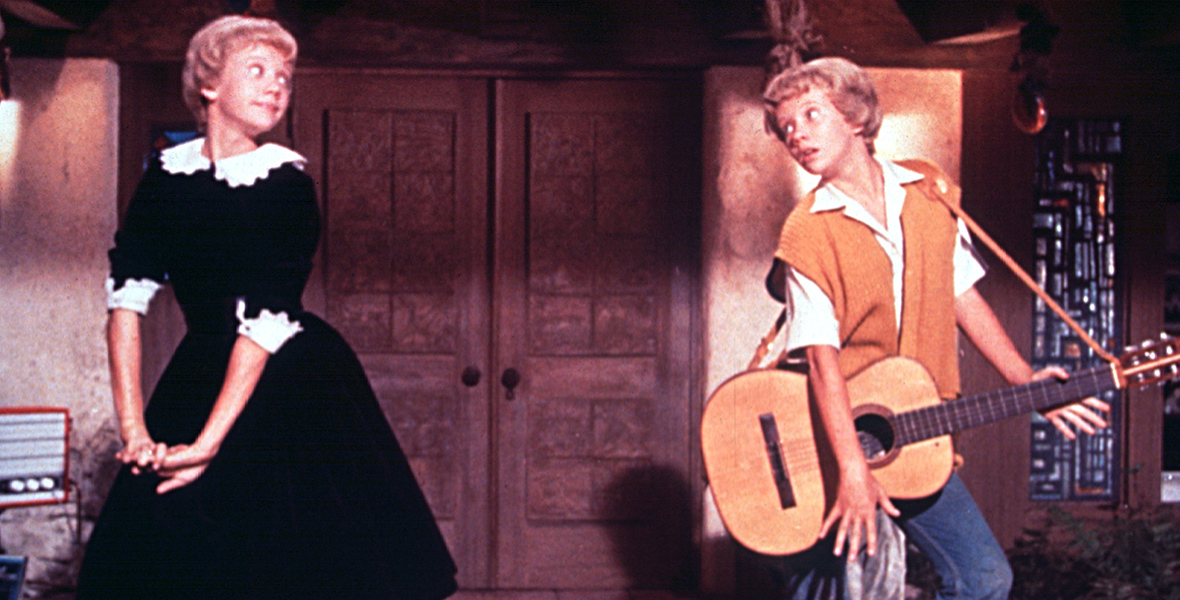 In a still from the 1961 Parent Trap, twins Susan and Sharon stand onstage, facing each other, with the twin on the right playing guitar.