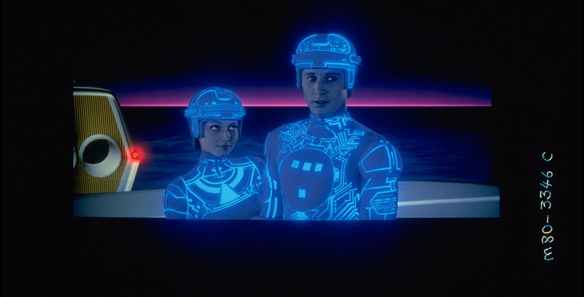 Yori and Tron, two humanoid computer programs in outfits with glowing blue lines, stand facing the camera. Yori is looking up at Tron, while Tron is looking at something off-screen.