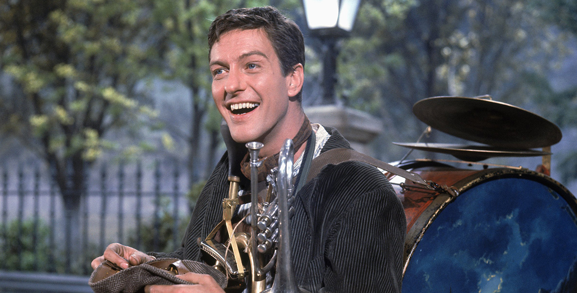 In a still from Mary Poppins, Bert smiles while holding his cap in his hands. He’s wearing a variety of instruments, including a drum on his back and a trumpet in his arms.