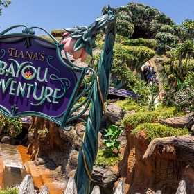 The lush, green exterior to the attraction Tiana’s Bayou Adventure. In front of the attraction is a sign stylized to appear held up by tall, blossoming flowers. The sign is purple and features the green logo for the attraction.