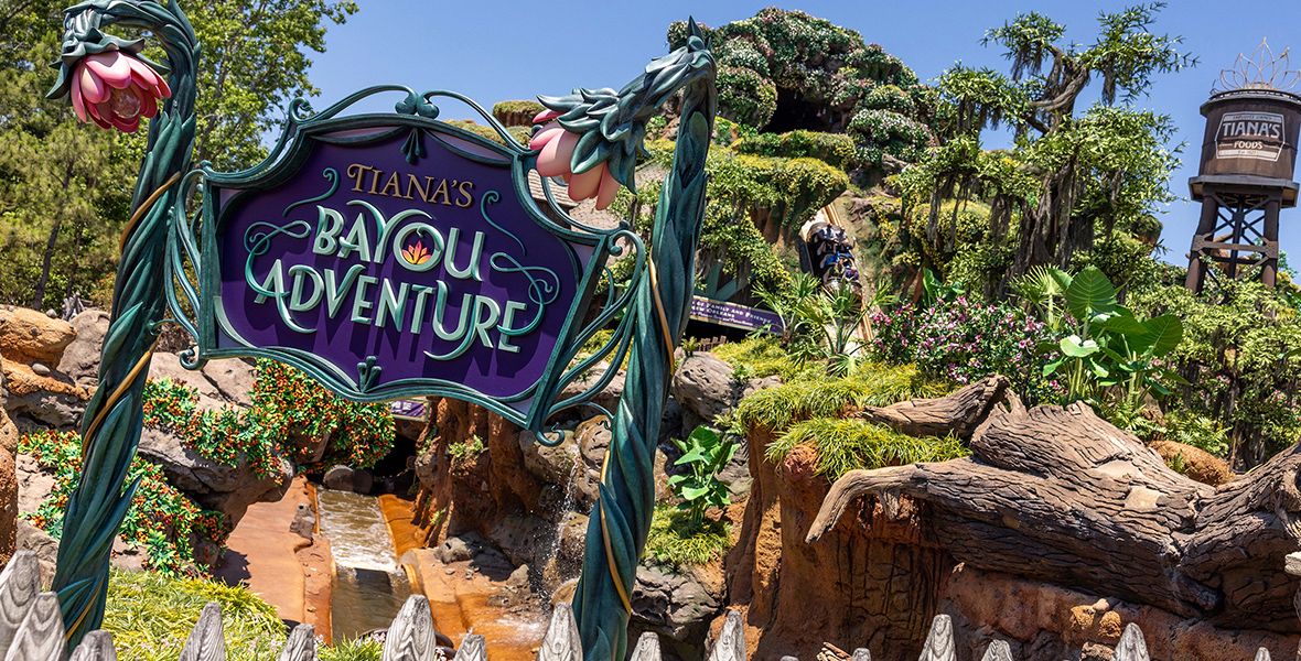 The lush, green exterior to the attraction Tiana’s Bayou Adventure. In front of the attraction is a sign stylized to appear held up by tall, blossoming flowers. The sign is purple and features the green logo for the attraction.
