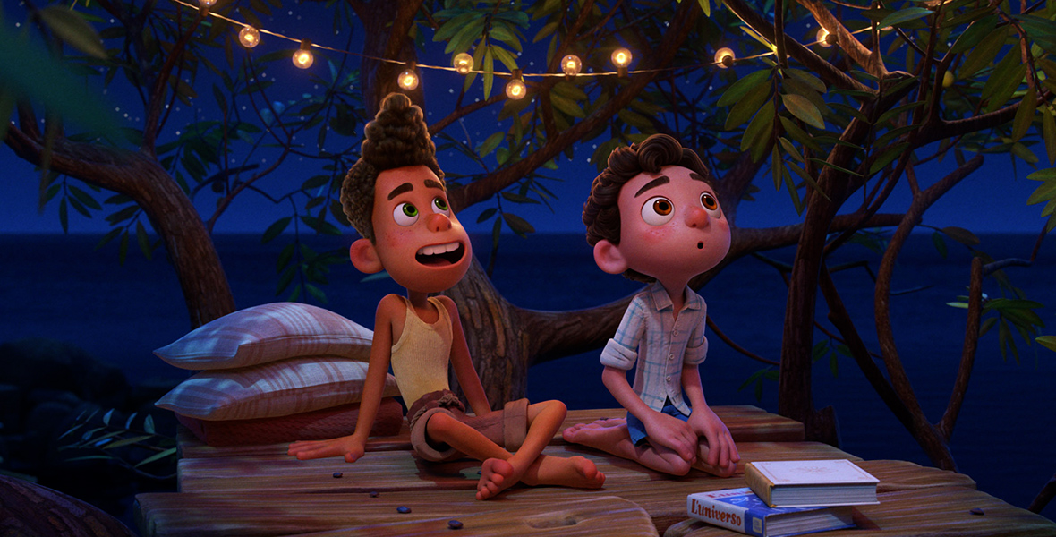 In a scene from the movie Luca, Italian boys Alberto (Jack Dylan Grazer), left, and Luca (voiced by Jacob Tremblay), right, are sitting on wooden planks built in a tree. Both gaze up at the stars, their expressions filled with wonder. Alberto sports a yellow tunic top paired with brown cargo pants, while Luca wears a white checkered flannel top with blue streaks and blue Bermuda shorts. String lights gracefully drape across branches, casting a soft glow. The backdrop showcases a starry night sky overlooking the sea.