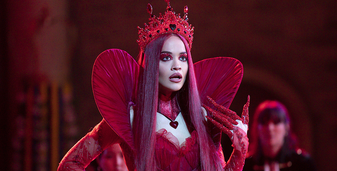 In a scene from the movie Descendants: The Rise of Red, Rita Ora as the Queen of Hearts wears a blood-red satin gown. The gown features a round collar adorned with ruched fabric in the same deep red hue, creating a heart-shaped illusion rising from her shoulders. Around her neck, she wears a paneled neck cuff with a large, heart-shaped pendant dangling from it. Completing her ensemble is a blood-red crown adorned with black hearts, and she has straight, raven-red hair. She wears sheer red lace gloves embellished with beads and sequins—and her left arm, lifted up, is adorned with long finger gloves sparkling with red stones.