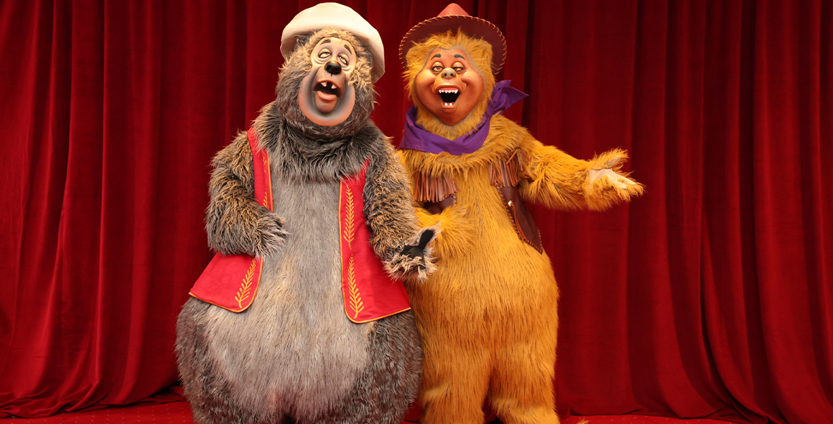 Big Al and Wendell debut costumes for Country Bear Musical Jamboree: an stitched vest and a handkerchief. They are standing on a red carpet in front of a red curtain.