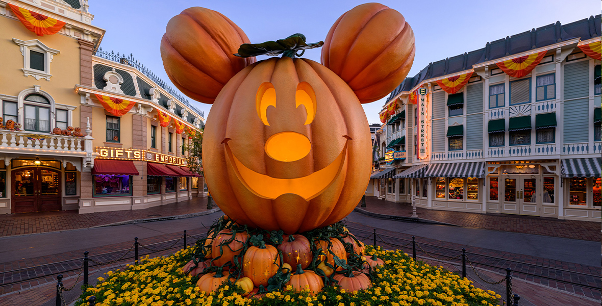 A giant, smiling Mickey Mouse pumpkin in the center of Disneyland Park’s Main Street U.S.A. sits on smaller pumpkins, surrounded by yellow flowers. Black bollards connected with chains encircle the display. In the background, shops and restaurants line the street with signs reading “Gift Emporium,” “Main Street,” and “Magic.”