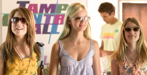 In a scene from Aquamarine, Hailey Rogers (JoJo), Aquamarine (Sara Paxton), and Claire Brown (Emma Roberts) are standing next to each other. Hailey is wearing an orange tank top with green butterflies and dark-tinted sunglasses. Aquamarine is wearing a purple tank top and pink sunglasses. Claire is wearing a pink top with ruffles and flowers patterns and gray sunglasses.