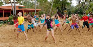 In a scene from Teen Beach Movie, Brady (Ross Lynch) and McKenzie (Maia Mitchell) dance with other actors on a sandy beach. Brady wears a short-sleeve yellow top and blue bathing suit shorts with orange stripes. McKenzie sports a black zip-up top with pink accents, paired with patterned bathing shorts. Palm trees and an orange building with a white rood create a backdrop.